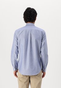 Camisa de botones azul claro, con tela texturizada y un patrón sutil, de mangas largas y dobladillo redondeado, combinada con pantalones beige.