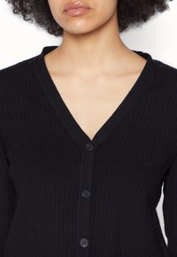 Cardigan negro de canalé con escote en V, que presenta seis botones negros en la parte delantera. Textura suave, con mangas largas y un ajuste ceñido.