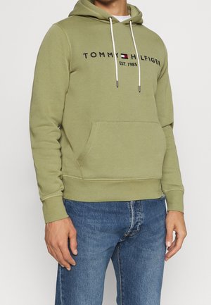 Homme portant un sweat à capuche Tommy Hilfiger vert olive avec poche avant, cordons blancs, et un jean bleu, debout devant un fond uni.