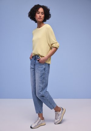 Kvinde med krøllet hår iført en lys gul trøje, blå jeans og sølvfarvede sneakers, stående foran en ensfarvet lyseblå baggrund.