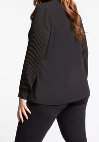 Svart långärmad blus med ribbad textur på ärmarna, avslappnad passform, rund nederkant och sidofickor, i kombination med mörka byxor.