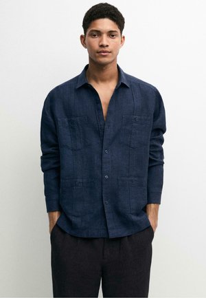 Junger Mann trägt ein dunkelblaues Hemd mit Knöpfen und vier Fronttaschen, steht vor einem schlichten hellen Hintergrund, Hände in den Taschen.