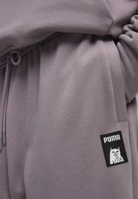 Fioletowe dresy wykonane z miękkiego materiału, z regulowanym ściągaczem w talii oraz czarną prostokątną naszywką z logo kota i napisem "PUMA".