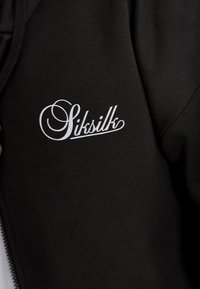 Čierna mikina na zips s textúrovanou tkaninou, s výrazným bielym vyšitým logom "SikSilk" na ľavej strane hrudníka.