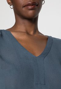 Haut en V bleu avec un tissu texturé, présentant des coutures subtiles et une coupe décontractée, mettant en valeur un décolleté lisse et des contours définis.