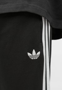 Čierne športové nohavice s bielym dizajnom troch pásikov, s malým bielym vyšívaným logom Adidas blízko pásu. Hladká textúra látky.