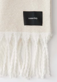 Witte, getextureerde sjaal met franjes. Bevat een zwart label met het woord "someday." Zacht fabric met subtiele patronen.