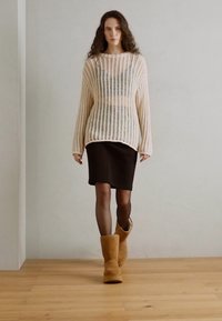 Suéter de malha creme com corte largo e mangas longas, combinado com uma saia escura e botas de camurça até ao joelho em tom bege, sobre um chão de madeira.