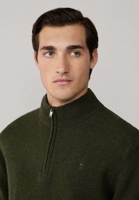 Suéter de color verde oliva con cremallera en el cuello, en una textura de punto, que presenta un alto cuello y un pequeño emblema en el pecho, fabricado con una suave mezcla de lana.
