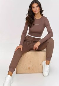 Brązowy prążkowany crop top o długim rękawie z logo na piersi, w połączeniu z dopasowanymi dresami ze sznurkiem i białymi sneakersami, siedzący na drewnianym bloku.