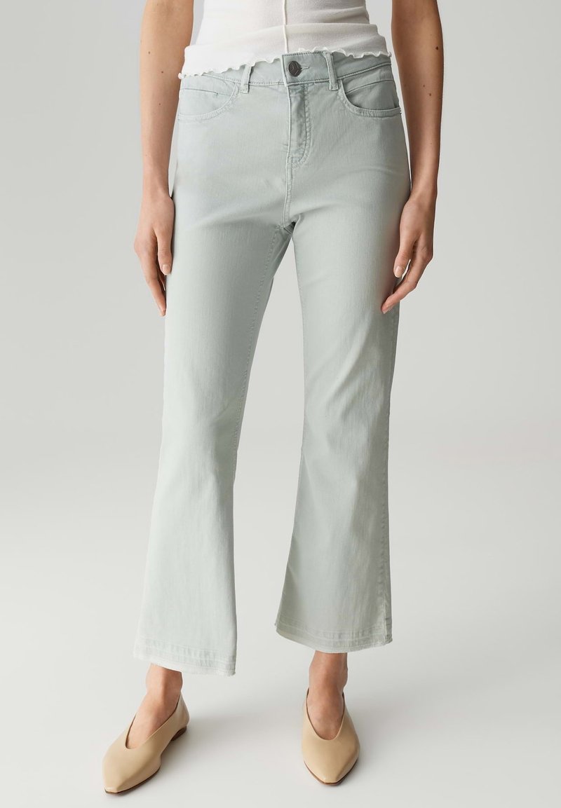 Jeans verde chiaro a palazzo con una texture liscia, design a cinque tasche, chiusura frontale con bottone e dettagli di cucitura discreti. Abbinati a scarpe beige.