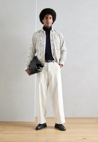 Camisa ligera con estampado en crema y diseño de paisley azul, cuello alto negro, pantalones blancos holgados y zapatos oscuros. Sosteniendo un bolso negro.