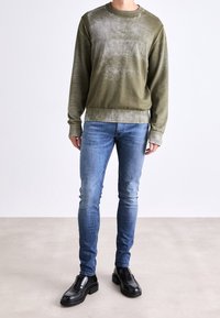 Sudadera verde oliva con acabado texturizado, combinada con jeans ajustados azul y mocasines de cuero negros. Tela suave y puños acanalados.