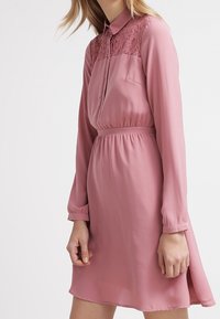 Langærmet pink kjole med blonder detaljer på skulderpartiet, kraven og et elastisk bånd i livet for en tætsiddende silhuet. Glat stoftekstur.