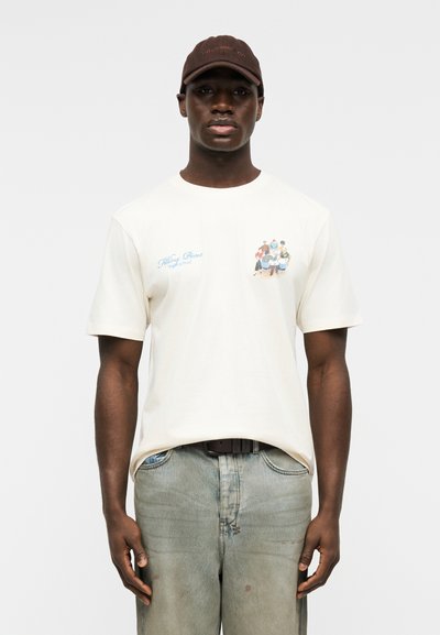 Άνδρας με σκούρο δέρμα, φοράει καφέ καπέλο, εκρού μπλουζάκι με μικρό πολύχρωμο σχέδιο και κείμενο, και ξεθωριασμένο ανοιχτό μπλε τζιν, στέκεται μπροστά σε απλό φόντο.