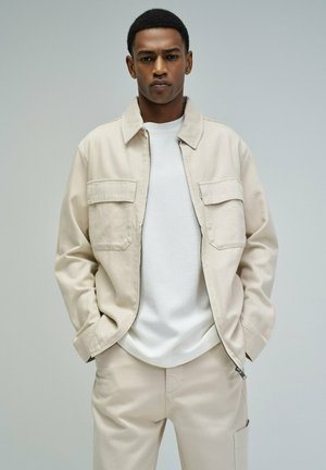 Joven con chaqueta y pantalones beige y camisa blanca, de pie frente a un fondo liso gris claro.