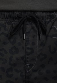 Gros plan d'une ceinture à cordon noire sur un pantalon gris foncé avec un motif léopard subtil et des coutures visibles.