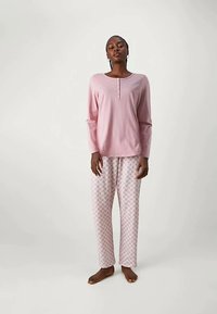 Kvinna stående barfota i en långärmad rosa topp och ljusa mönstrade pyjamasbyxor mot en enfärgad bakgrund.