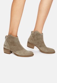 Botas de tobillo de ante gris con perforaciones recortadas, tacón bajo y apilado, y cremallera lateral para el cierre. Textura suave con diseño de punta redondeada.
