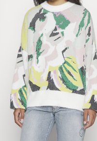 Pletený svetr s barevným abstraktním vzorem v zelené, růžové, žluté a šedé barvě, s načechraným bílým lemem a volným střihem.