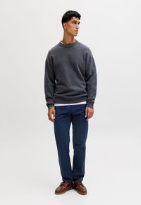 Ung mand står og kigger mod højre, iført en grå sweater, marineblå bukser og brune lædersko på en ensartet lys baggrund.