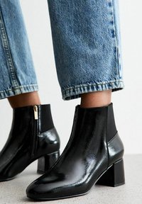 Botines negros de charol con tacón cuadrado, con paneles laterales elásticos y cremallera lateral. Llevados con unos vaqueros claros de mezclilla recortados.