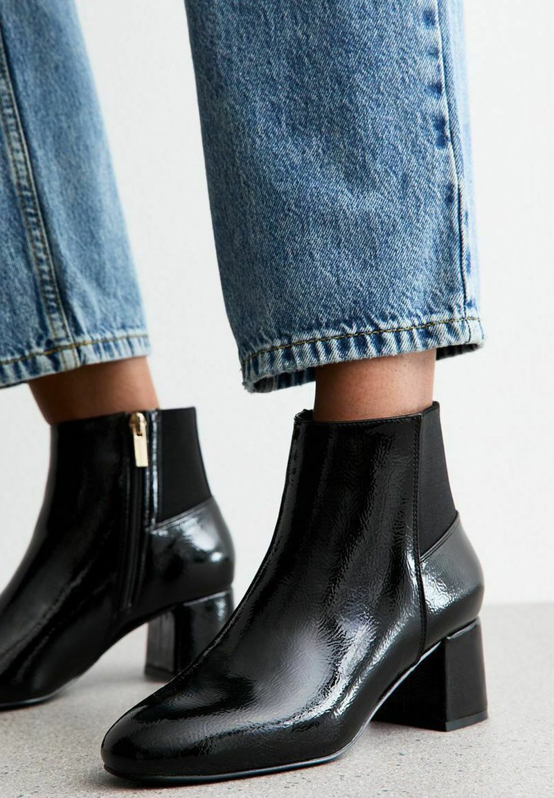 Botines negros de charol con tacón cuadrado, con paneles laterales elásticos y cremallera lateral. Llevados con unos vaqueros claros de mezclilla recortados.