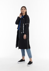 Cappotto lungo nero con cerniere, dotato di una tasca a toppa con la scritta "LOVE", indossato sopra una maglietta blu e jeans chiari con risvoltini, abbinato a sneakers nere.
