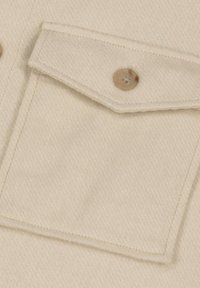 Beige stofclose-up met een borstzak met een klep, beveiligd door een ronde, lichtbruine knop met vier gaten. Zachte textuur en egale kleur.