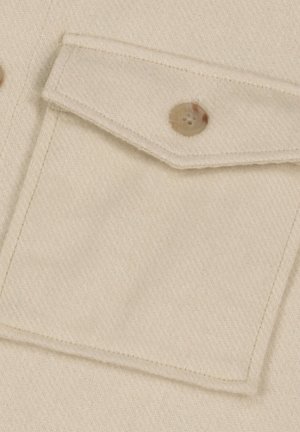 Beige stofclose-up met een borstzak met een klep, beveiligd door een ronde, lichtbruine knop met vier gaten. Zachte textuur en egale kleur.