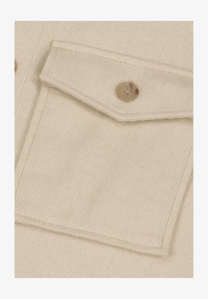 Primo piano su un tessuto beige con una tasca sul petto dotata di alette, fissata da un bottone rotondo di colore marrone chiaro con quattro fori. Tessuto morbido e colore uniforme.