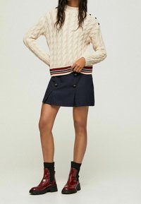 Cremfarbener Strickpullover mit einem gestreiften roten und marineblauen Saum, kombiniert mit einem marineblauen, plissierten Minirock mit goldenen Knöpfen. Schwarze Knöchelschäfte.