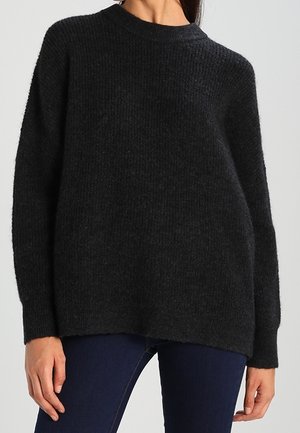 Femme portant un pull en maille noir ample à manches longues associé à un jean bleu foncé sur un fond uni.