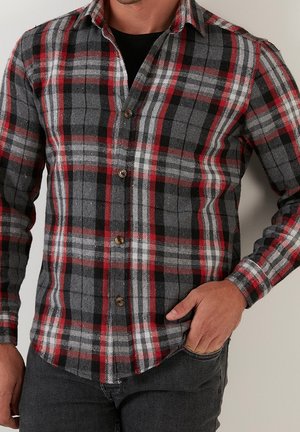 Uomo che indossa una camicia a quadri rossa, nera, bianca e grigia con bottoni e una maglietta nera sotto, mano in tasca, su uno sfondo neutro.