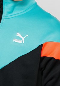 Chaqueta Puma con sección superior en turquesa, sección inferior negra y detalle naranja, confeccionada en tela texturizada con cierre de cremallera.