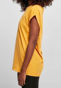 Keltainen puuvillapaita, jossa on lyhyet käännetyt hihansuut, rento istuvuus, pyöreä pääntie ja sileä pinta. Helma on hieman pidempi takana.