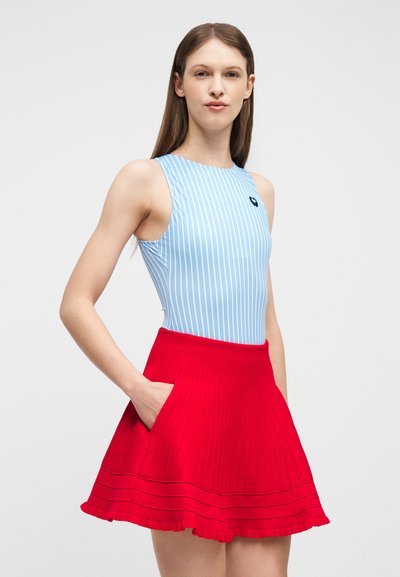 Jeune femme portant un haut sans manches à rayures bleues et blanches et une jupe plissée rouge avec des poches, debout devant un fond uni clair.