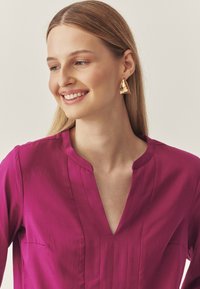 Blusa magenta con scollatura a V e pieghe verticali, abbinata a grandi orecchini d'oro con un design astratto. Tessuto dalla texture liscia.