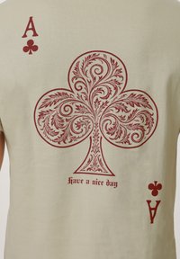 Πίσω μέρος μπεζ πουκάμισου με μεγάλο διακοσμητικό κόκκινο σύμβολο μπαστούνι, κόκκινο γράμμα "A" και εικονίδια χαρτιών μπαστούνι στις γωνίες, καθώς και το κείμενο «καλή σου μέρα» κάτω από το μπαστούνι.