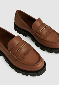 Brune læder loafers med en fronttassel-detalje og en guldfarvet accent, der har en tekstureret sort gummisål. Glat overfladefinish.
