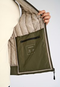 Olivegrüne Puffermantel-Innenseite mit hellem Futter, Reißverschlusstasche und Pflegeetikett; durchgehend steppte Textur; wasserabweisende Eigenschaften.