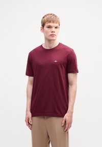 Jonge man in een effen bordeauxrood T-shirt en beige broek die tegen een witte achtergrond staat en iets naar de zijkant kijkt.