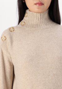 Maglione beige con colletto a costine, dotato di cinque grandi bottoni chiari sulla spalla. Tessuto morbido, vestibilità comoda.