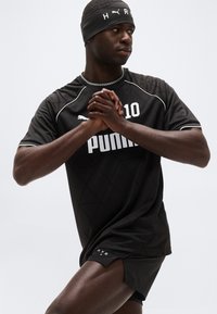 Αθλητής με μαύρα αθλητικά ρούχα Puma και κορδέλα στο κεφάλι, με τα χέρια ενωμένα, κοιτάζοντας προς τα πλάγια μπροστά σε ένα απλό φωτεινό φόντο.