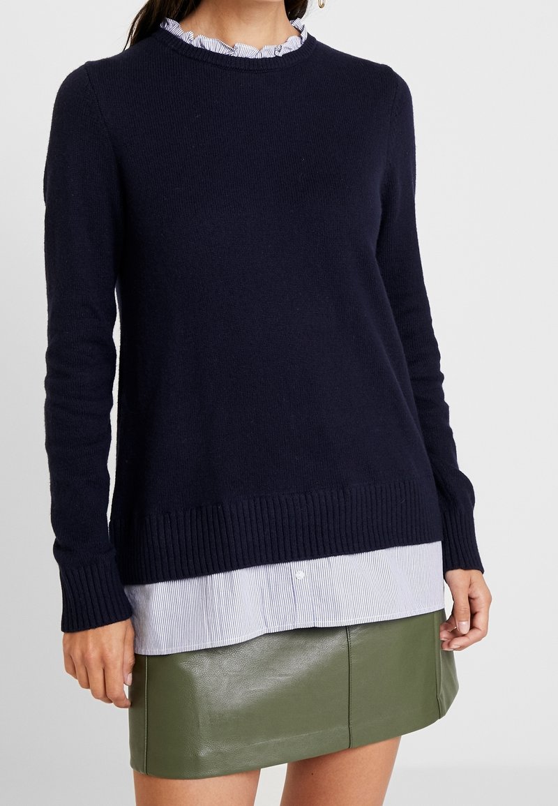 Pull en tricot bleu marine avec ourlet et poignets côtelés, superposé à une chemise à rayures bleues claire et blanches, associé à une jupe en cuir verte.