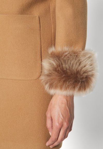 Cappotto di lana cammello con una grande tasca frontale e bordo in pelliccia sintetica marrone chiaro sulle maniche, caratterizzato da una texture liscia e una vestibilità sartoriale.