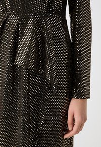 Robe noire avec des pois métalliques dorés, manches longues et détail péplum à la taille, une main reposant à côté du corps.