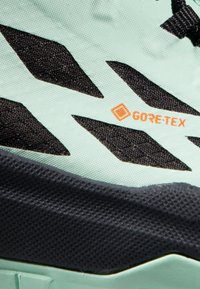 Scarpe da trail di colore verde chiaro con accenti neri, caratterizzate da una superficie testurizzata, un tallone rinforzato e un logo Gore-Tex arancione in evidenza.