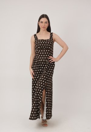 Mujer vestida con un vestido maxi marrón sin mangas con lunares blancos y una abertura frontal, de pie con la mano en la cadera contra un fondo liso.