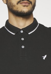 Svart polo-skjorte med tre knapper, hvit kant på kragen, og en liten hvit fuglelogo på venstre bryst. Strukturert stoff.